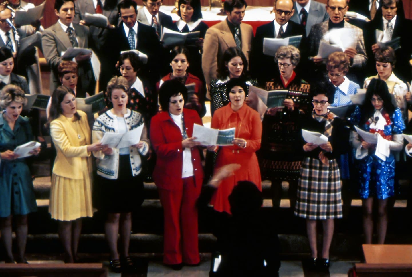 Un coro canta partituras en plataformas escalonadas dirigido por un director.